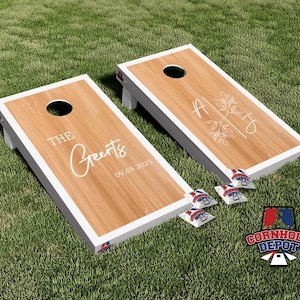 Może przedstawiać: Dwie drewniane deski do cornhole z białymi obramowaniami i otworem u góry. Jedna deska ma napis "The Greerts" i datę "09.05.2025". Druga ma inicjały i kwiatowy wzór. Zawiera worki z fasolą.
