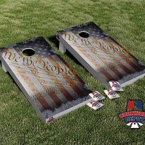 May include: Two wooden cornhole boards with an American flag design and the text "We the People." The boards have a hole in the top and are accompanied by red, white, and blue bean bags. A logo for Cornhole Depot is visible.