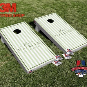 Può includere: Due tavole da cornhole rettangolari con un motivo a righe verticali verde chiaro e bianco. Ogni tavola presenta un foro circolare e il testo "the WALKERS" con la data "06 13 2026" stampata in caratteri scuri. Sono inclusi quattro sacchetti.
