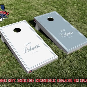 May include: Two rectangular cornhole boards, one white and one light blue, each featuring a circular hole at the top, a double-line border, and the text "THE Palmers 10.18.26" printed in white or blue. The boards have white side supports.