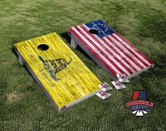 May include: Two wooden cornhole boards with a distressed wood look. One board features a yellow background with a Gadsden flag design and the text "Don't Tread On Me". The other board features a red, white, and blue American flag with 13 stars in a circle. The boards are on a green grassy surface.
