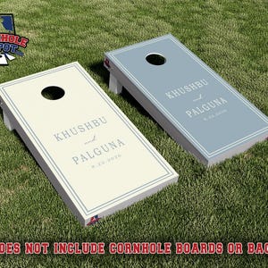 May include: Two rectangular cornhole boards with circular holes, one cream and one slate blue. Both feature a white border and the printed text KHUSHBU and PALGUNA 8.22.2026. The boards are angled on white legs, displaying a custom design.