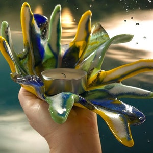 Puede incluir: Un portavelas de vidrio con forma de mano, colorido, con tonos amarillos, azules, verdes y blancos. Contiene una peque&ntilde;a vela de t&eacute; encendida. El vidrio tiene un dise&ntilde;o texturizado y fluido, creando una pieza decorativa &uacute;nica.