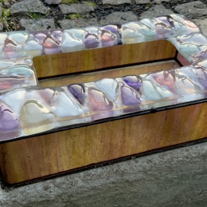 Könnte beinhalten: Ein rechteckiger Taschentuchbox-Deckel aus Glas mit einem Mosaikdesign aus schillernden violetten, weißen und klaren Glasstücken. Die Box hat einen Holzsockel mit einer braunen Holzmaserung.