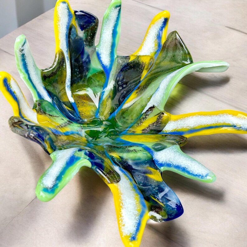 Puede incluir: Un cuenco de vidrio abstracto y colorido con un dise&ntilde;o &uacute;nico de estrella. El cuenco presenta tonos vibrantes de amarillo, verde, azul y blanco, creando un efecto visual llamativo. El di&aacute;metro del cuenco es de aproximadamente 30 cm.