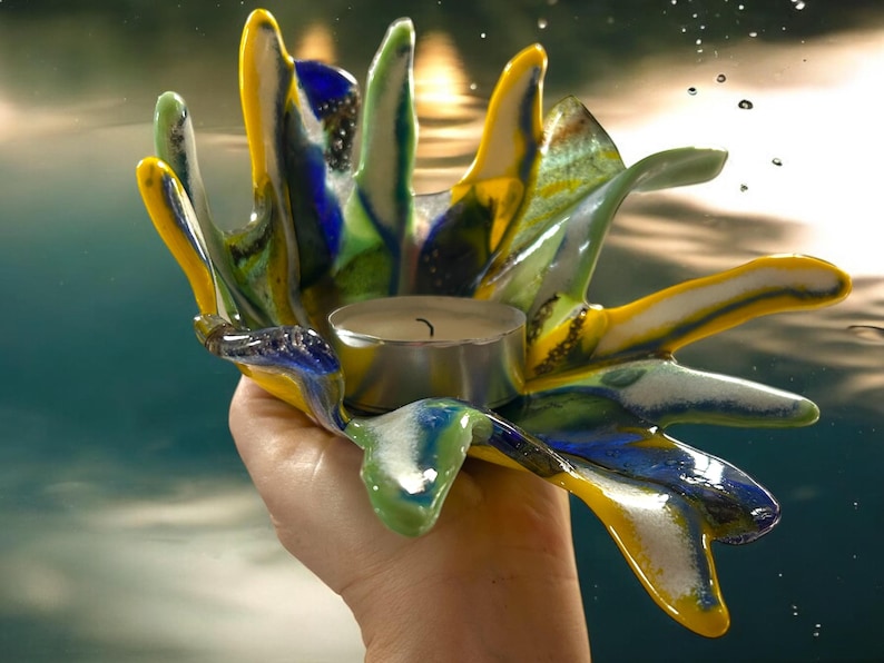 Puede incluir: Un portavelas de vidrio con forma de mano, colorido, con tonos amarillos, azules, verdes y blancos. Contiene una peque&ntilde;a vela de t&eacute; encendida. El vidrio tiene un dise&ntilde;o texturizado y fluido, creando una pieza decorativa &uacute;nica.