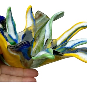 Puede incluir: Escultura abstracta de vidrio con colores vibrantes. La pieza presenta tonos de amarillo, azul, verde y blanco, creando un dise&ntilde;o din&aacute;mico. La forma de la escultura recuerda a una flor floreciendo o una explosi&oacute;n de energ&iacute;a. Medidas en cent&iacute;metros.