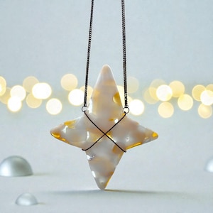 Puede incluir: Un colgante en forma de estrella con un diseño veteado blanco y amarillo, suspendido por una cadena oscura. La estrella está hecha de un material translúcido, y la cadena está sujeta con una cruz de alambre. El fondo tiene luces borrosas de tonos cálidos.