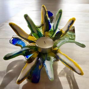 Puede incluir: Un portavelas de vidrio artesanal y colorido. El portavelas presenta un dise&ntilde;o de estrella con tonos vibrantes de amarillo, azul, verde y blanco. Una copa de metal plateado sostiene una vela de t&eacute; blanca encendida. El vidrio tiene una apariencia texturizada y org&aacute;nica.