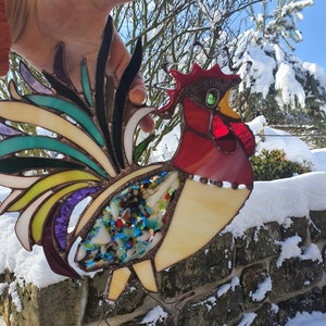 Puede incluir: Un gallo de vidrio coloreado con una cola multicolor y una cabeza roja. El gallo está hecho de vidrio y metal y es una pieza decorativa.