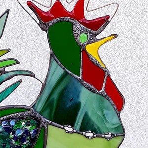 May include: A stained glass rooster with a red comb, green body, and yellow beak. The rooster is made of small pieces of glass that are joined together with lead. The rooster is set against a white background.