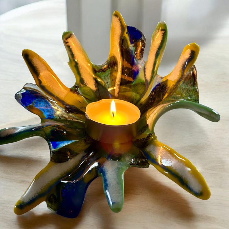 Puede incluir: Un portavelas de vidrio artesanal y colorido con un dise&ntilde;o abstracto vibrante. El portavelas presenta un hueco central para una vela de t&eacute;, rodeado de formas radiantes en forma de p&eacute;talos en tonos amarillos, verdes, azules y blancos. La vela est&aacute; encendida.