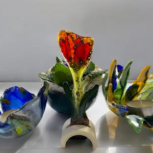 Puede incluir: Colecci&oacute;n de coloridas piezas de arte en vidrio. La pieza central es una escultura de flor de vidrio rojo y amarillo. A los lados, dos candelabros de vidrio en tonos azules, verdes y amarillos. Una peque&ntilde;a planta en maceta en el fondo.