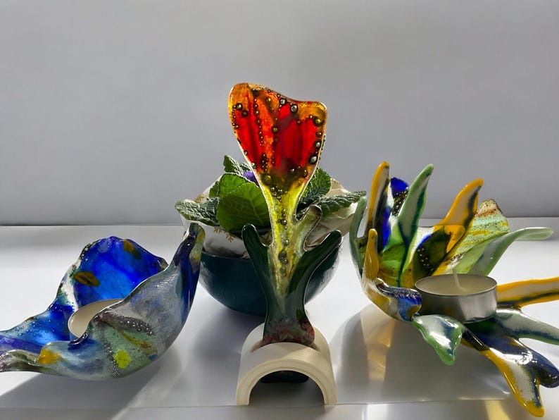 Puede incluir: Colecci&oacute;n de coloridas piezas de arte en vidrio. La pieza central es una escultura de flor de vidrio rojo y amarillo. A los lados, dos candelabros de vidrio en tonos azules, verdes y amarillos. Una peque&ntilde;a planta en maceta en el fondo.