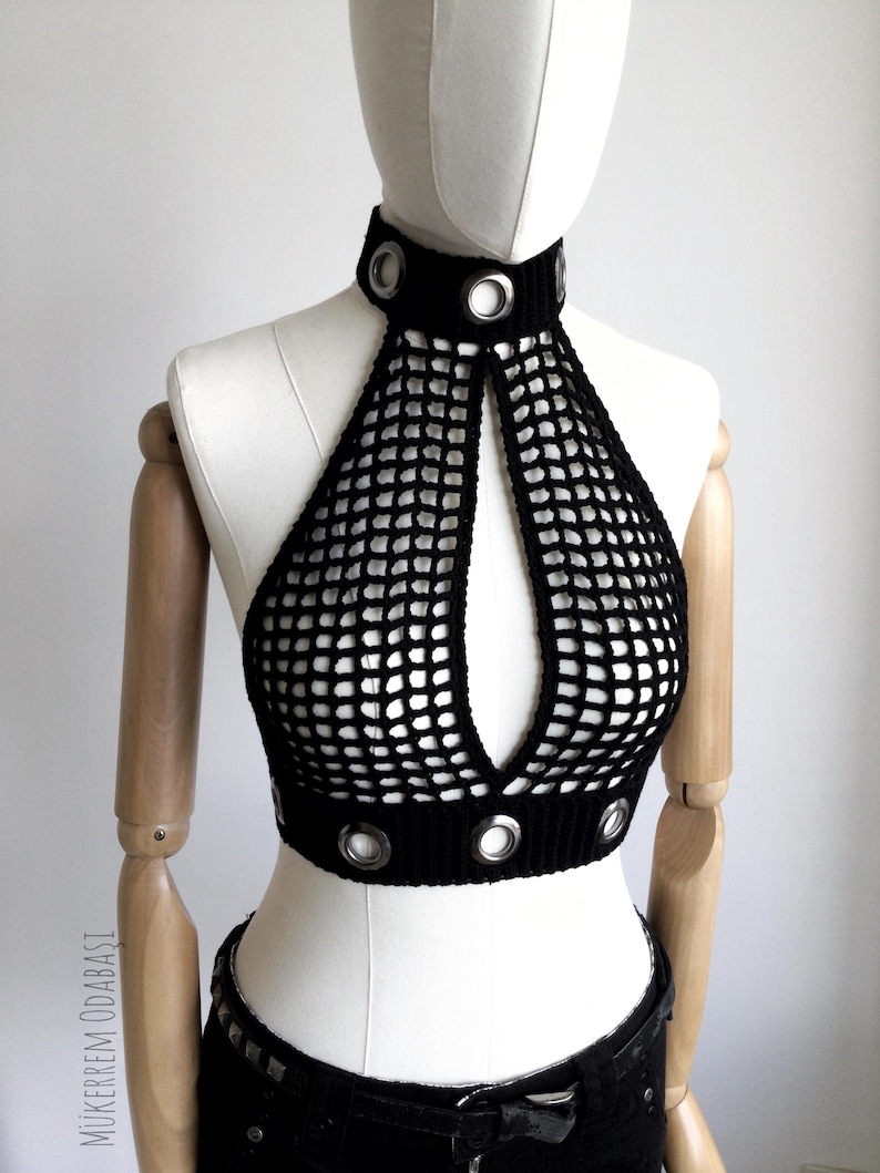 May include: Black crochet halter top with a high collar and a keyhole design. The top features silver grommets around the collar and the bottom hem. The top is displayed on a mannequin.