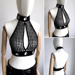 May include: Black crochet halter top with a high collar and a keyhole design. The top features silver grommets around the collar and the bottom hem. The open weave creates a net-like appearance. The top is displayed on a mannequin.