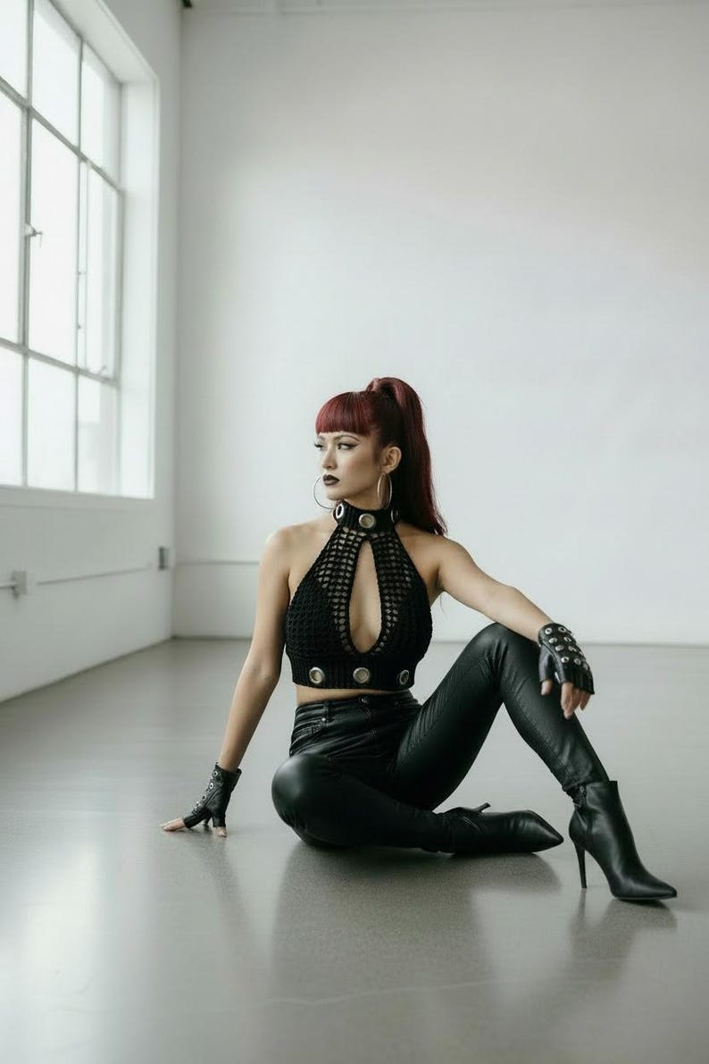 May include: A black halter top with a keyhole design and silver accents. The top is paired with black leather pants and boots. The model is wearing fingerless gloves and hoop earrings. The background is a white studio setting.