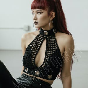 May include: Black crochet halter top with a keyhole neckline and silver grommet details. The top is part of a fashion ensemble. The model has red hair and is wearing large hoop earrings.