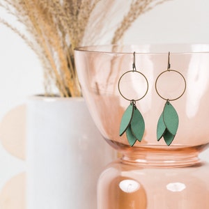 May include: A pair of green leather leaf earrings with gold hoops. The earrings are hanging from a pink glass bowl.