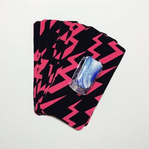 Peut inclure: Un jeu de cartes avec un motif de foudre noir et rose. Les cartes sont &eacute;tal&eacute;es en &eacute;ventail et un objet d&eacute;coratif bleu et blanc est plac&eacute; sur les cartes.