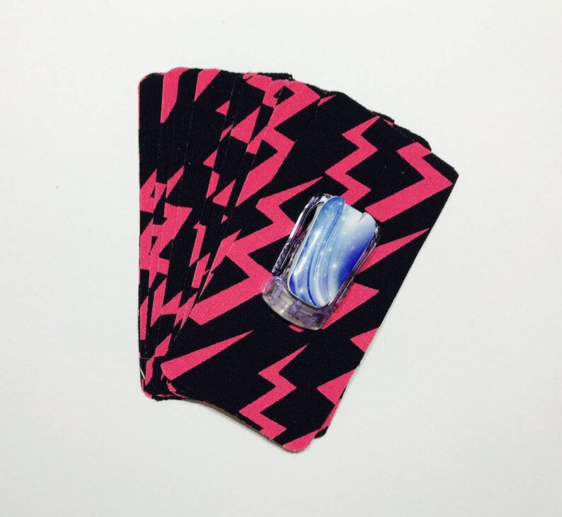 Peut inclure: Un jeu de cartes avec un motif de foudre noir et rose. Les cartes sont &eacute;tal&eacute;es en &eacute;ventail et un objet d&eacute;coratif bleu et blanc est plac&eacute; sur les cartes.