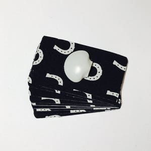 Op de afbeelding: Een stapel zwarte stofvierkanten met witte hoefijzerpatronen. Een witte plastic knop is bevestigd aan het bovenste vierkant.