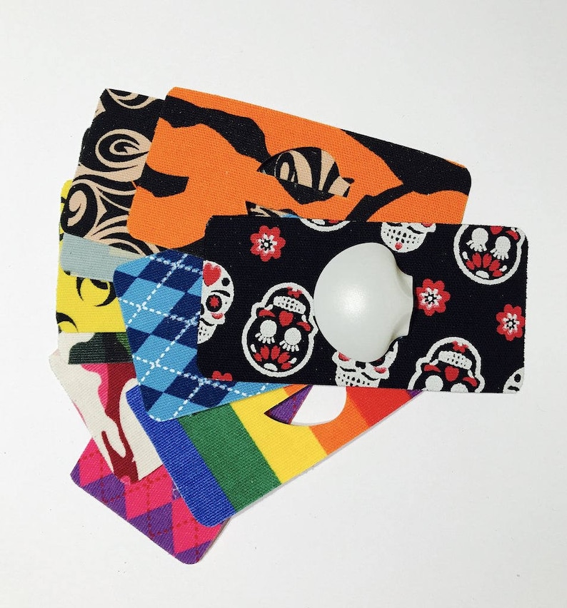 Op de afbeelding: Een stapel kleurrijke stofstalen met verschillende patronen, waaronder een zwart-wit schedelontwerp, een regenboogstreep en een blauw-wit ruitpatroon. De stalen zijn in een waaier vorm gerangschikt, met het bovenste staal met een witte knop.