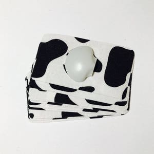 Op de afbeelding: Een stapel witte stoffen onderzetters met een zwart koe-print ontwerp. Een witte ronde knop ligt bovenop de stapel.