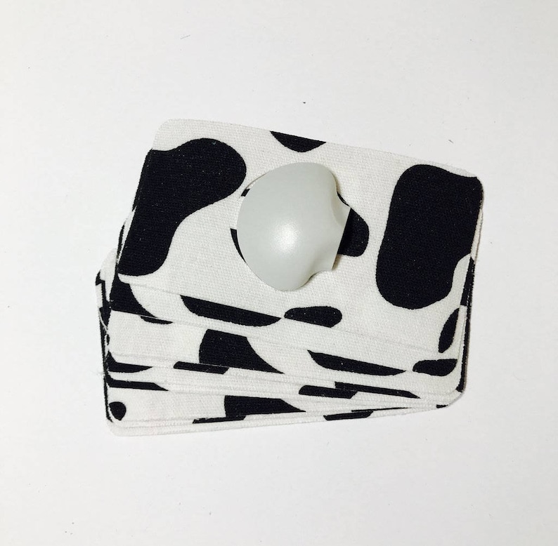 Op de afbeelding: Een stapel witte stoffen onderzetters met een zwart koe-print ontwerp. Een witte ronde knop ligt bovenop de stapel.