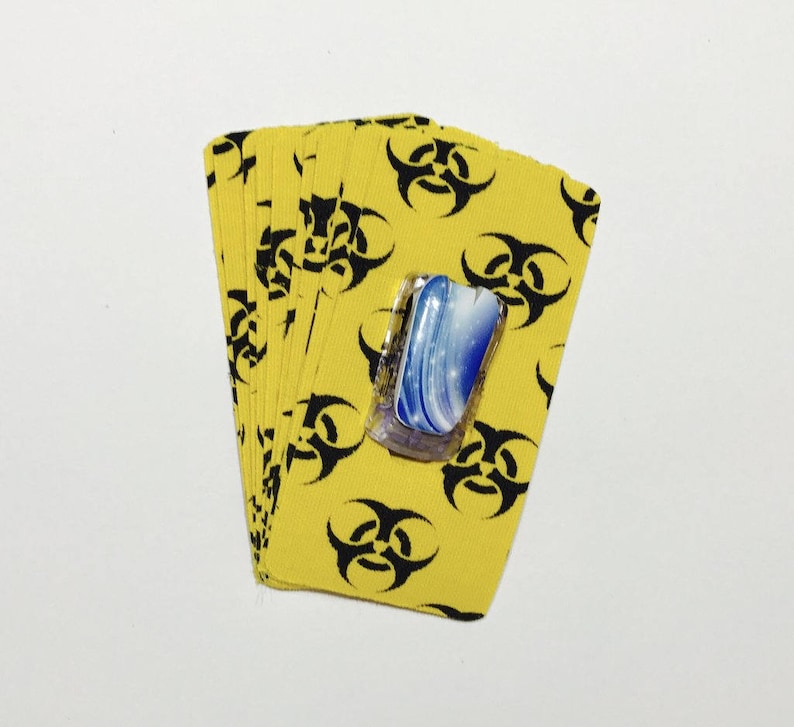Peut inclure: Un jeu de cartes avec un fond jaune et des symboles de danger biologique noirs. Les cartes sont &eacute;tal&eacute;es en &eacute;ventail et une petite tuile de verre bleu et blanc est pos&eacute;e sur le dessus du jeu.