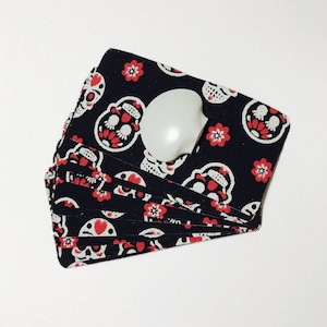 Op de afbeelding: Een stapel zwarte stoffen kaarten met een patroon van witte schedels en rode bloemen. De kaarten zijn dubbelgevouwen en hebben een witte plastic knop aan de bovenkant.