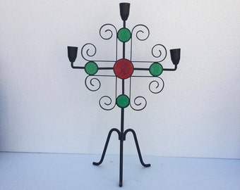 May include: Black metal candelabra with a cross-shaped design and five candle holders. The cross is decorated with green and red glass circles.