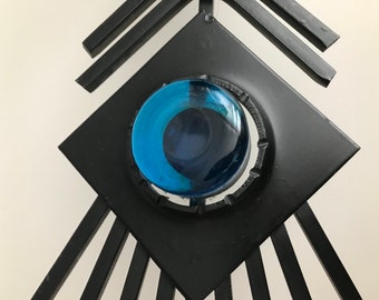 May include: A black metal wall sculpture with a blue glass circle in the center. The sculpture is in the shape of a diamond with radiating lines.