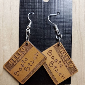 May include: A pair of wooden earrings with the text "Hello my name is Basic B*tch" on each earring. The earrings are brown and have a silver hook.