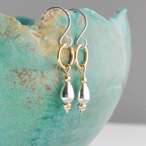 May include: A pair of silver teardrop earrings with gold accents. The earrings are hanging from a ceramic bowl.