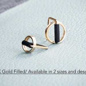 May include: A pair of gold-filled earrings with a circular design and a black bar in the center. The earrings are displayed on a light blue surface. Text on the image reads: 14K Gold Filled/ Available in 2 sizes and designs.