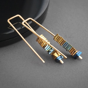 May include: A pair of gold earrings with a geometric design. Each earring features a long, thin gold bar with a stack of small, square-shaped beads in shades of blue and gold.