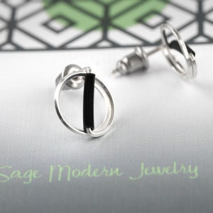 May include: A pair of silver stud earrings with a circular design and a vertical black bar. The earrings are displayed on a white card with the text "Sage Modern Jewelry" in green. The background features a geometric pattern.