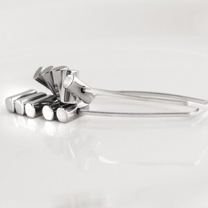 May include: A pair of silver-colored tongs with a unique design. The tongs feature a series of rectangular blocks on one end and a curved handle. The design is modern and minimalist, suitable for serving food or as a decorative item.