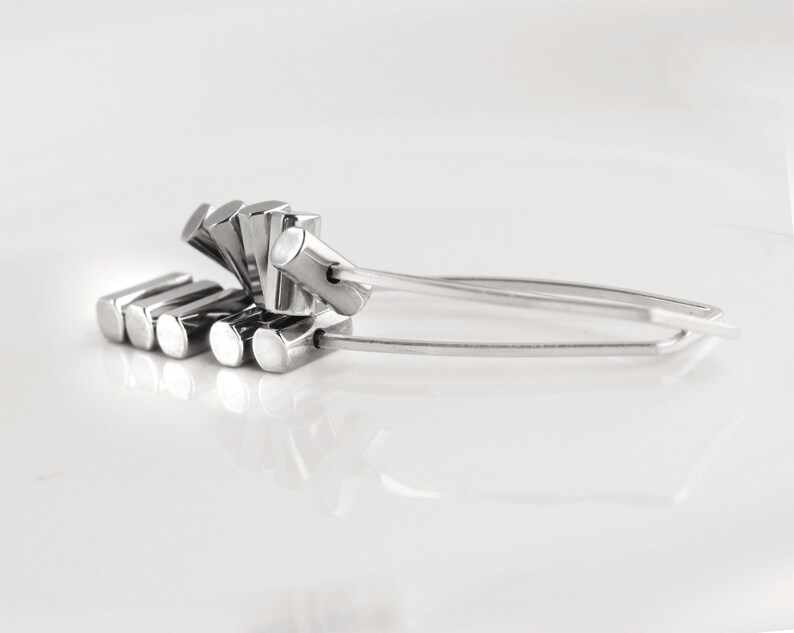 May include: A pair of silver-colored tongs with a unique design. The tongs feature a series of rectangular blocks on one end and a curved handle. The design is modern and minimalist, suitable for serving food or as a decorative item.