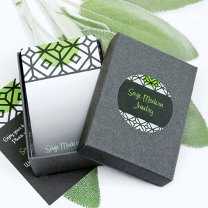 May include: Open gray jewelry box with a geometric design in green and white. The box is labeled "Sage Modern Jewelry" in green text. Sage green leaves are in the background. A small card with the text "Sage Modern" is visible.