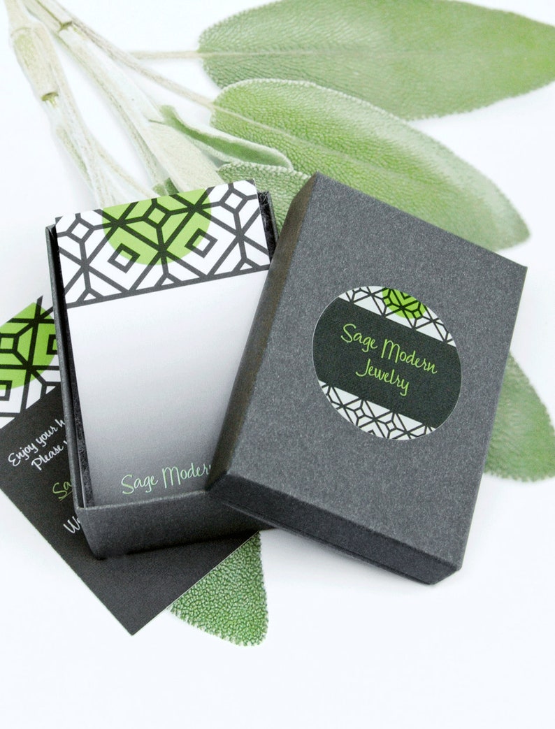 May include: Open gray jewelry box with a geometric design in green and white. The box is labeled "Sage Modern Jewelry" in green text. Sage green leaves are in the background. A small card with the text "Sage Modern" is visible.