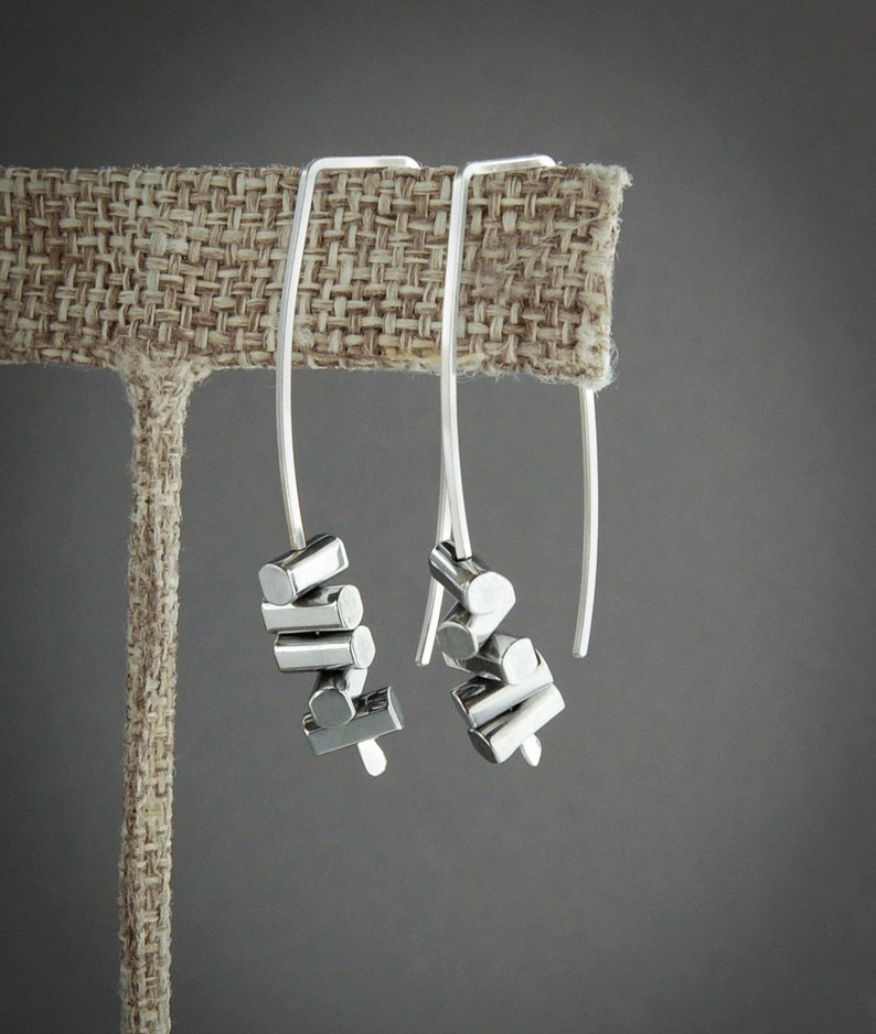 May include: A pair of silver drop earrings. Each earring features a long, curved wire with a cluster of small, cylindrical silver beads. The earrings are displayed on a neutral-toned fabric background against a gray backdrop.