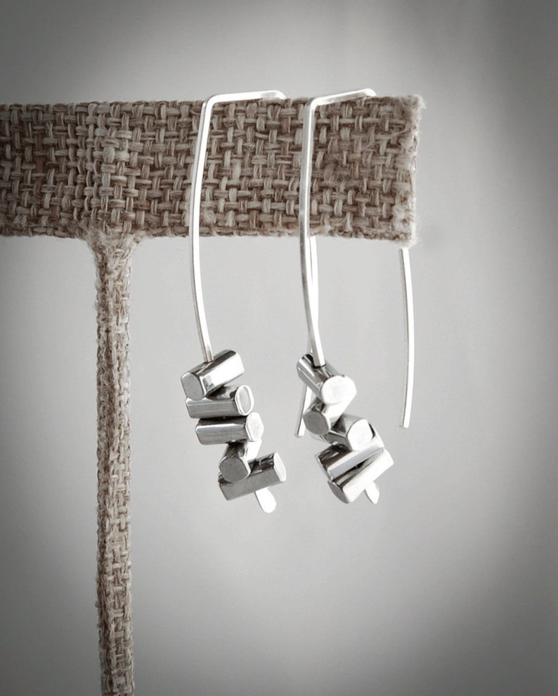 May include: Two silver dangle earrings with geometric shapes. The earrings are hanging from a brown fabric display stand.