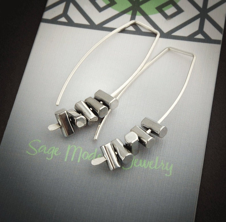 May include: A pair of silver earrings with a modern design. Each earring features a long, curved silver wire with a cluster of small, geometric, silver-colored beads. The earrings are displayed on a gray and white patterned surface.