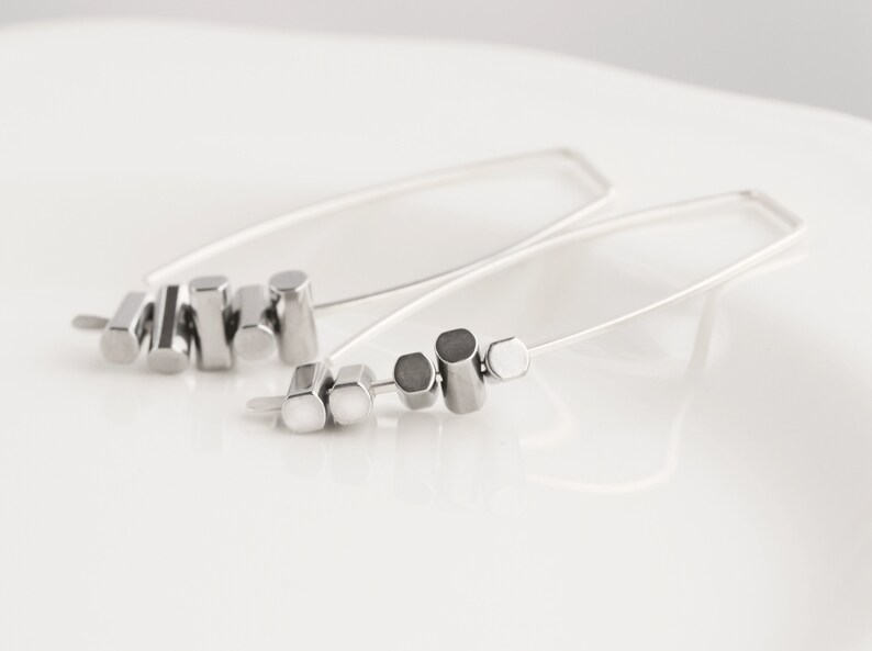 May include: A pair of silver-toned earrings. Each earring features a long, thin, curved wire with a series of geometric shapes, including cylinders and hexagons. The earrings are displayed on a white surface.