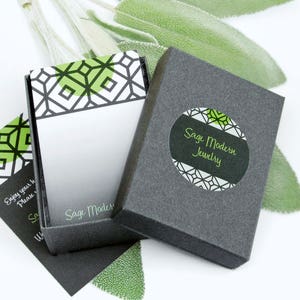 Peut inclure: Une bo&icirc;te &agrave; bijoux noire avec un motif g&eacute;om&eacute;trique vert et blanc sur le couvercle. La bo&icirc;te est ouverte et une petite carte avec le texte "Sage Modern Jewelry" est visible &agrave; l'int&eacute;rieur. La bo&icirc;te est pos&eacute;e sur une surface blanche.
