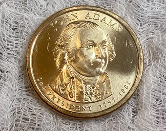 Op de afbeelding: Een gouden Amerikaanse dollarmunt met een portret van John Adams, de tweede president van de Verenigde Staten. De munt heeft de tekst "John Adams" boven het portret en "2nd President 1797-1801" eronder.