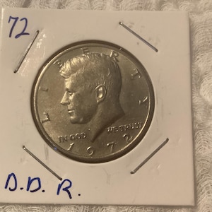 May include: A silver US half dollar coin featuring a profile of John F. Kennedy with the inscription "IN GOD WE TRUST" and the year "1972".