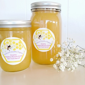 May include: Two glass jars of honey with silver lids. The jars are labeled "Premium Quality Organic Amazing Grapefruit Blossom Honey 2021 Fresh Harvest". A bouquet of white baby's breath flowers is in the foreground.